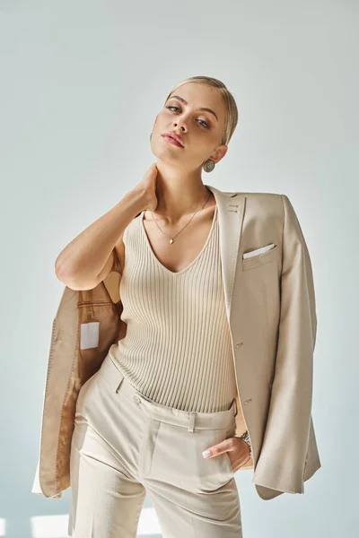 Sinnliches Mode-Model in eleganter beiger Kleidung, die den Hals berührt und mit der Hand in der Tasche auf grau posiert — Stockfoto