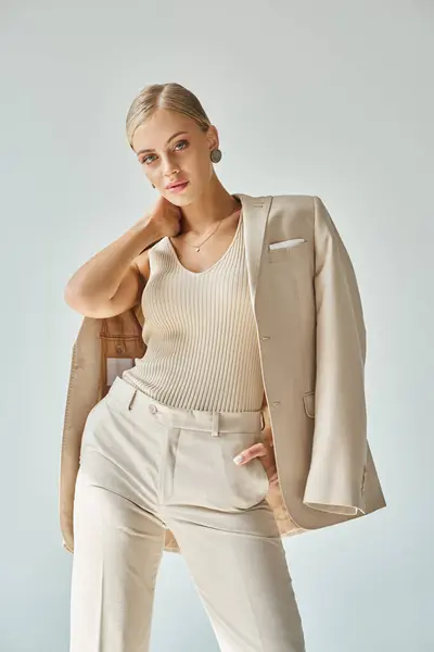 Modelo de moda joven en ropa beige con estilo tocando el cuello y posando con la mano en el bolsillo en gris - foto de stock