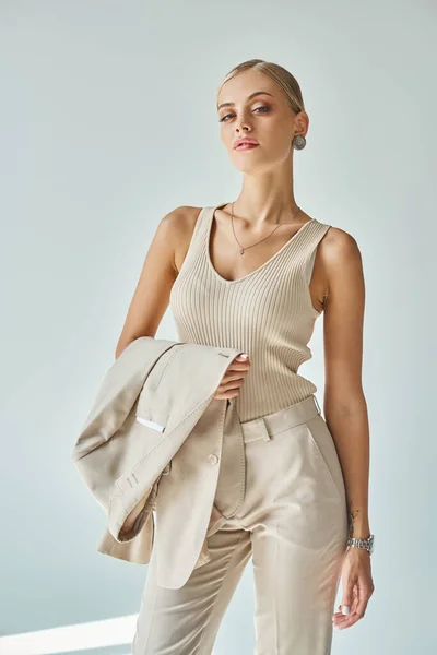 Mujer glamour en camiseta sin mangas beige sosteniendo chaqueta y mirando a la cámara en gris, belleza y moda - foto de stock