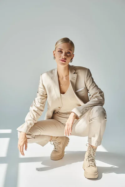 Trendy blonde woman in beige suit and laced boots posing on haunches on grey backdrop with sunlight — Stock Photo