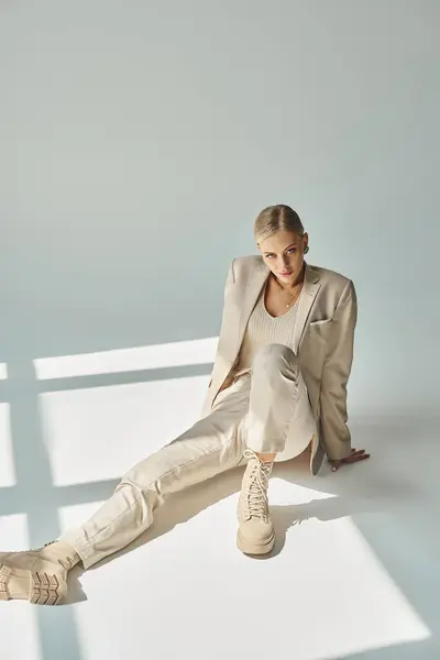 Charming woman in total beige outfit sitting in sunlight and looking at camera on grey, full length — Stock Photo