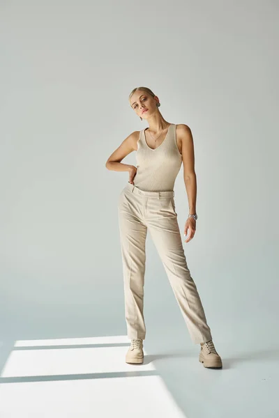 Full length of young fashionista in beige tank top and pants posing with hand on hip on grey — Stock Photo