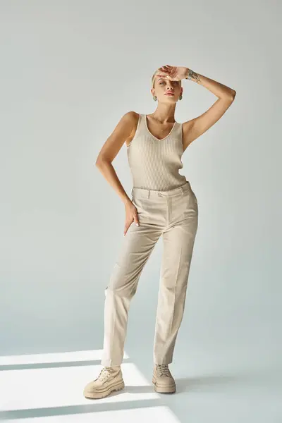Stylish blonde woman in tank top and pants posing with hand near forehead on grey, fashion shoot — Stock Photo