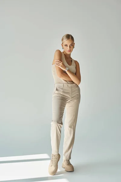 Blonde woman in pants and tank top standing with folded arms on grey with sunlight, urban style — Stock Photo