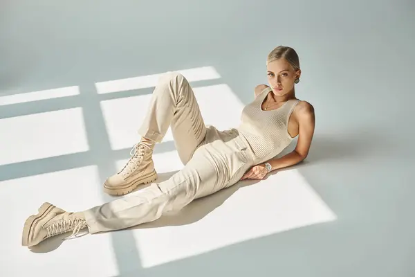Full length of stylish woman in tank top and pants posing in sunlight and looking at camera on grey — Stock Photo