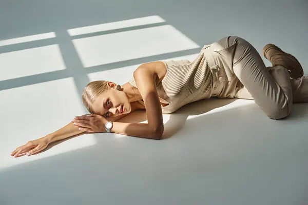 Alluring fashion model in total beige clothes lying down in sunlight and looking at camera on grey — Stock Photo