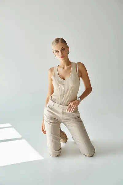 Trendy blonde woman in tank top and pants kneeling on grey backdrop with sunlight, urban fashion — Stock Photo