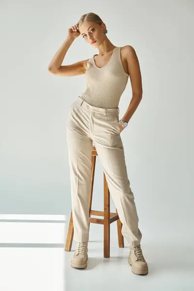 Full length of stylish woman posing with hand in pocket of beige pants near high stool on grey — Stock Photo