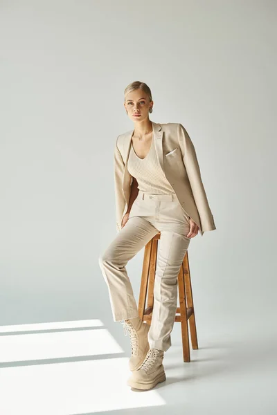 Elegant woman in beige suit and laced pants sitting on tall stool on grey backdrop with sunlight — Stock Photo