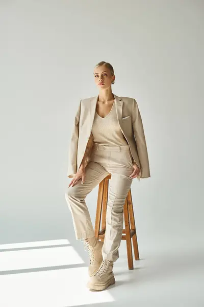 Dreamy young woman in pastel beige suit looking away on tall stool in sunlight on grey backdrop — Stock Photo