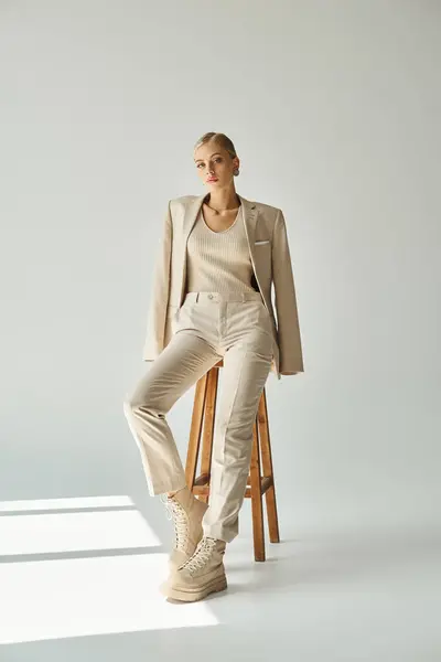 Pretty blonde woman in pastel beige suit looking at camera on tall stool in sunshine on grey — Stock Photo