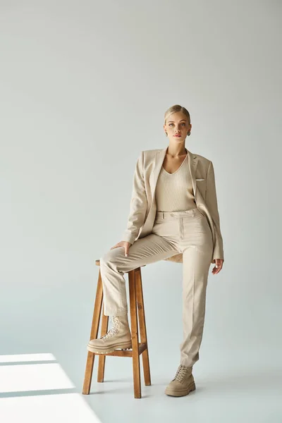 Young elegant woman looking at camera while posing with tall stool on grey backdrop with sunlight — Stock Photo