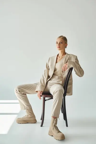 Modelo de moda rubia en traje beige pastel sentado en la silla y mirando hacia otro lado en el sol en gris - foto de stock