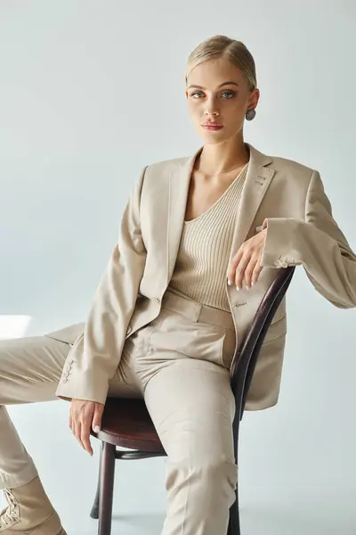 Blondes Mode-Model im pastellbeigen Anzug sitzt auf Stuhl und blickt vor grauem Hintergrund in die Kamera — Stockfoto