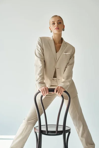 Séduisante mannequin de mode en costume beige pastel posant avec chaise et regardant la caméra sur fond gris — Photo de stock