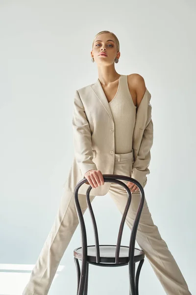 Attraktives weibliches Model im eleganten Anzug posiert mit Stuhl und blickt in die Kamera vor grauem Hintergrund — Stockfoto