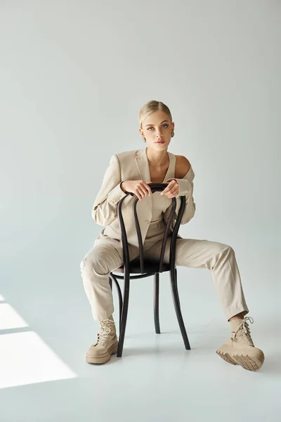 Junge Frau im beigen Outfit sitzt auf Stuhl und blickt in die Kamera auf grauen, urbanen Stil — Stockfoto