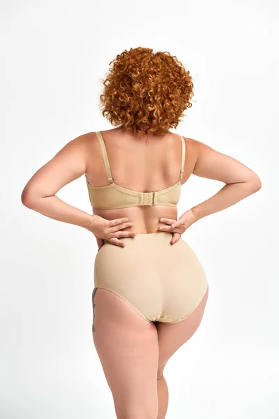 Plus size woman with red wavy hair posing in beige lingerie with hands on waist on white, back view — Stock Photo