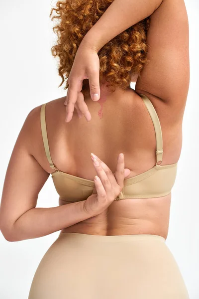 Back view of redhead woman in beige lingerie with hands behind back on white, plus size beauty — Stock Photo