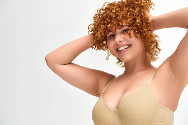 Cheerful young woman with curvy breasts touching red wavy hair on white backdrop, plus size beauty — Stock Photo