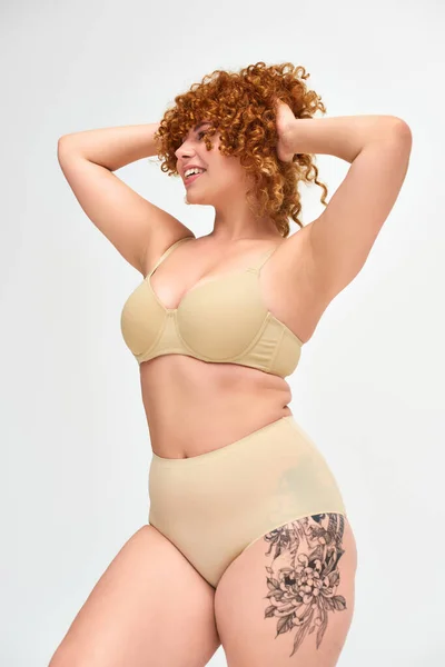 Donna curvy tatuata con capelli ricci rossi in posa in lingerie beige con le mani dietro la testa su bianco — Foto stock