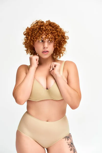 Découragé femme plus taille avec des cheveux bouclés rouges debout en lingerie beige et regardant loin sur blanc — Photo de stock