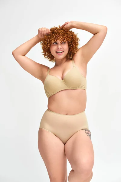 Overjoyed and redhead plus size model in beige underwear with raised hands looking away on white — Stock Photo