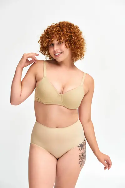 Flirty e smiley plus size modella in lingerie beige in posa con mano vicino alla spalla su bianco — Foto stock