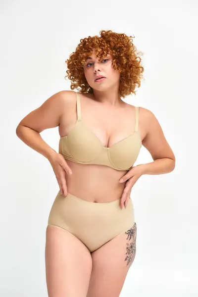 Sensual redhead plus size woman in beige lingerie posing with hands on waist on white, self-esteem — Stock Photo
