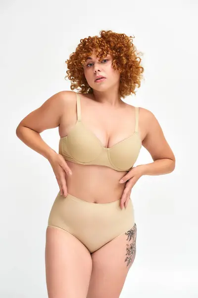 Sensual pelirroja mujer de talla grande en lencería beige posando con las manos en la cintura sobre blanco, autoestima - foto de stock