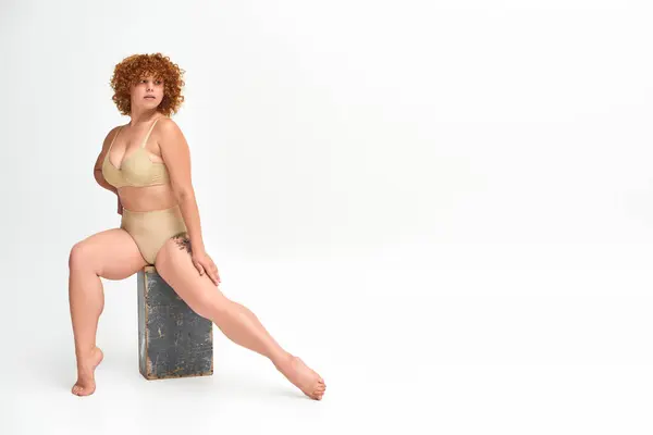 Longitud completa de la expresiva pelirroja más mujer de tamaño en lencería sentado en grunge caja de madera en blanco - foto de stock