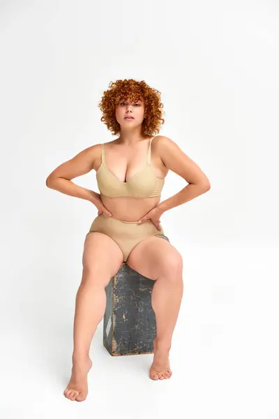 Redhead curvaceous woman in beige lingerie sitting on grunge wooden box with hands on waist on white — Stock Photo