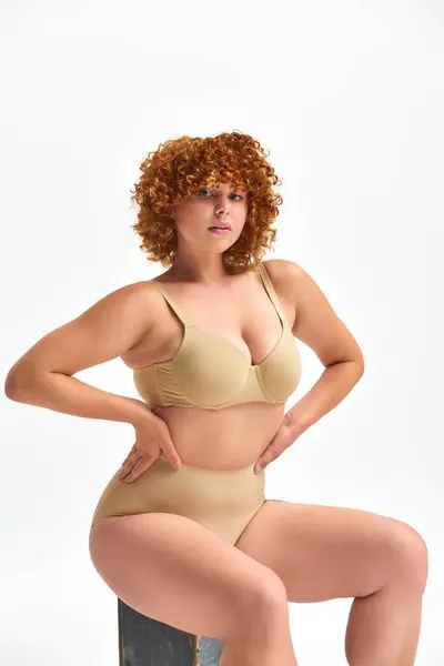Curvy redhead woman in beige lingerie sitting on wooden box with hands on waist on white backdrop — Stock Photo