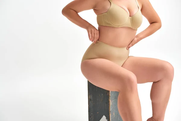 Corte vista de modelo feminino curvilíneo em lingerie sentado na caixa de madeira com as mãos na cintura em branco — Fotografia de Stock