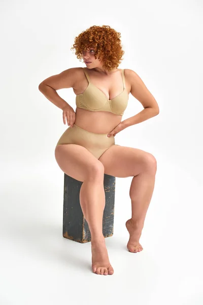 Redhead and curvaceous woman in beige lingerie sitting on wooden box and looking away on white — Stock Photo