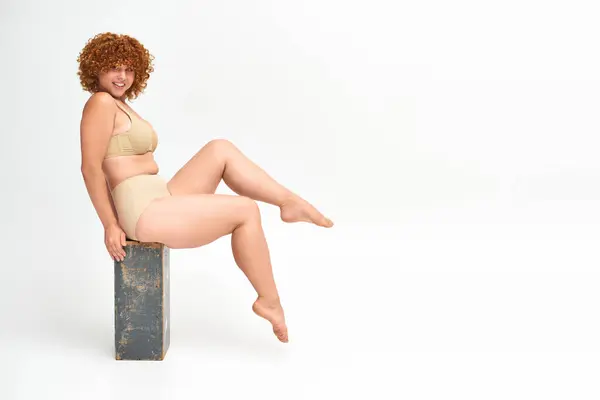 Eccitata e curvy rossa donna in lingerie beige seduta su scatola di legno grunge su bianco, lunghezza intera — Foto stock