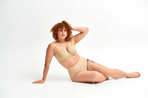 Alluring curvy woman in beige lingerie touching red wavy hair and looking at camera on white — Stock Photo
