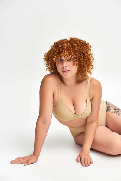 Seductive tattooed woman with red curly hair and plus size body looking at camera on white — Stock Photo