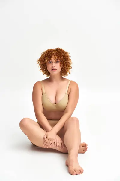 Full length of curvaceous redhead woman in beige lingerie sitting and looking at camera on white — Stock Photo