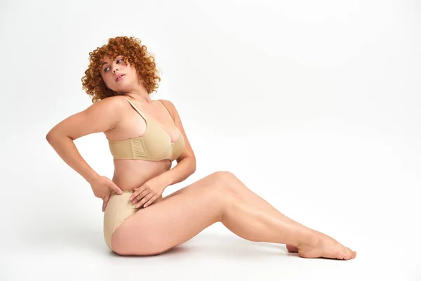 Full length of body positive redhead woman in beige lingerie looking away while sitting on white — Stock Photo
