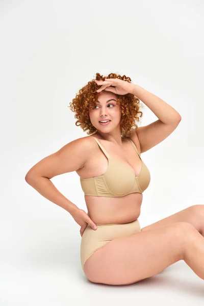 Smiling redhead plus size woman in beige lingerie sitting with hand on hip and looking away on white — Stock Photo