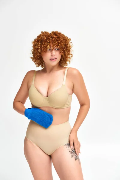 Redhead plus size woman in beige lingerie holding bath glove and looking away on white — Stock Photo