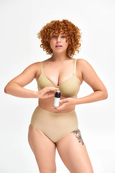 Young curvy woman in beige lingerie holding deodorant spray and looking at camera on white — Stock Photo
