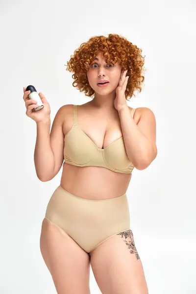 Amazed redhead plus size woman with roll-on deodorant touching face and looking at camera on white — Stock Photo