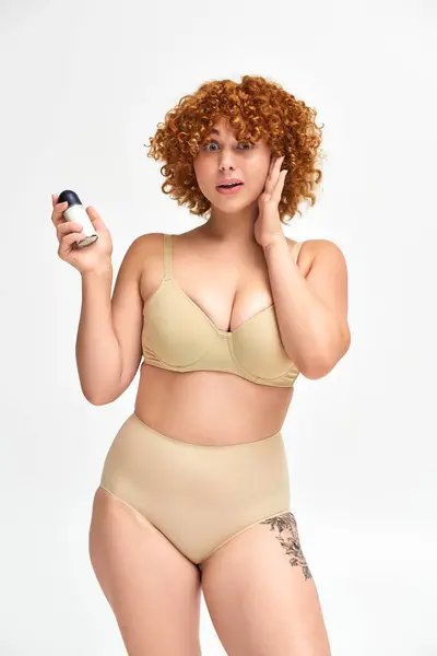 Rossa stupito plus size donna con roll-on deodorante toccare il viso e guardando la fotocamera su bianco — Foto stock