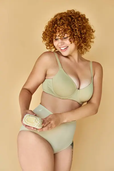 Pelirroja alegre más mujer de tamaño en ropa interior sosteniendo paño sobre fondo beige, mimos - foto de stock