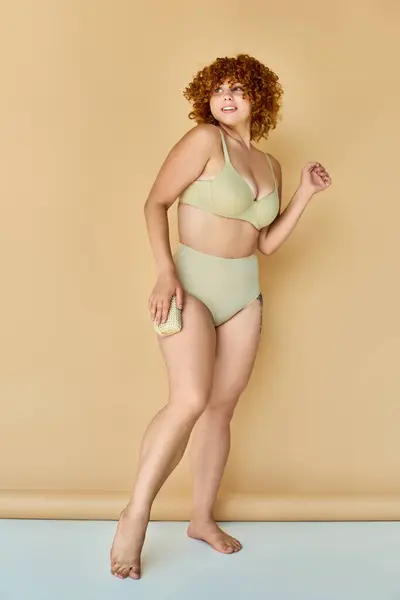 Piena lunghezza di donna rossa curvy in lingerie tenendo washcloth e guardando lontano sullo sfondo beige — Foto stock