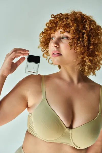 Encantador modelo curvilíneo en sujetador beige con pelo rojo sosteniendo botella de perfume y mirando hacia otro lado en gris - foto de stock