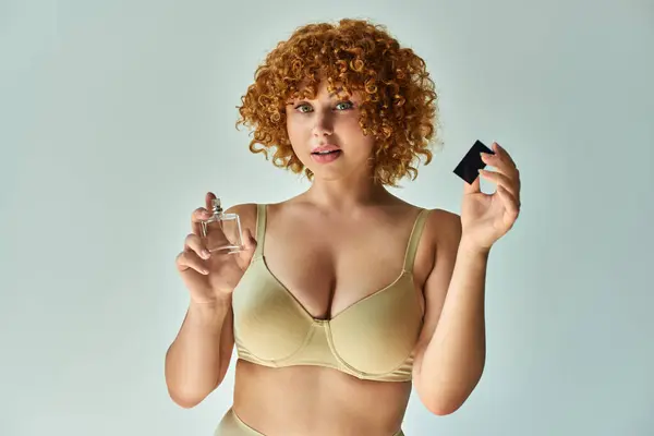 Sensual redhead woman in beige bra with curvy body spraying perfume and looking at camera on grey — Stock Photo