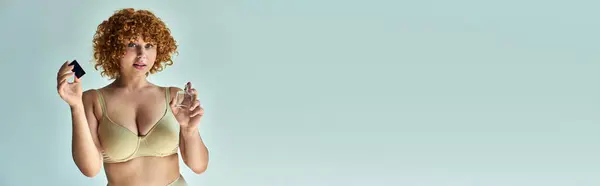 Seductora pelirroja en sujetador beige con perfume de pulverización corporal curvilínea sobre bandera gris horizontal - foto de stock